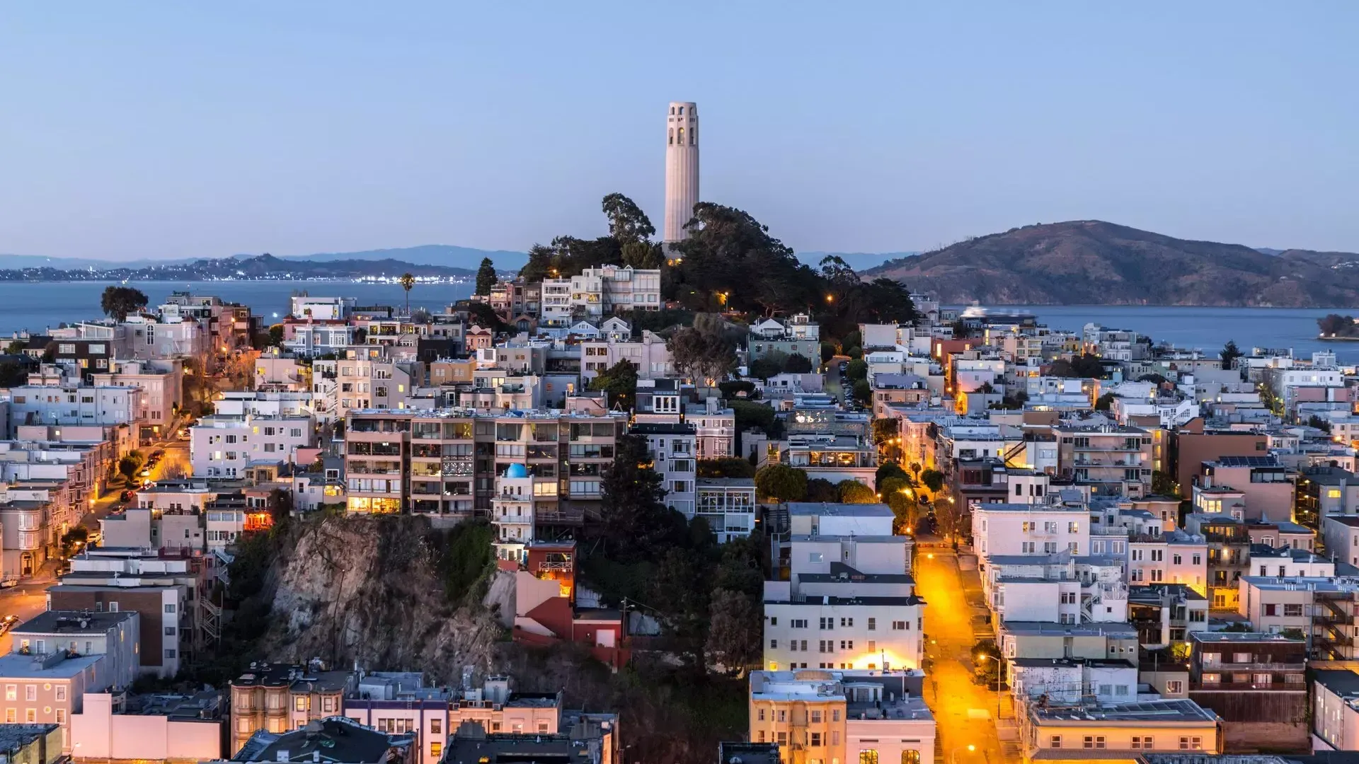 La Coit Tower de San Francisco au crépuscule, avec les rues éclairées devant elle et la baie de San Francisco derrière elle.